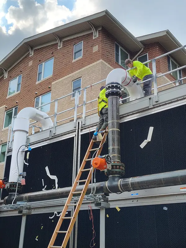 Crew fitting an insulated elbow at a rooftop cooling tower beside a brick facade